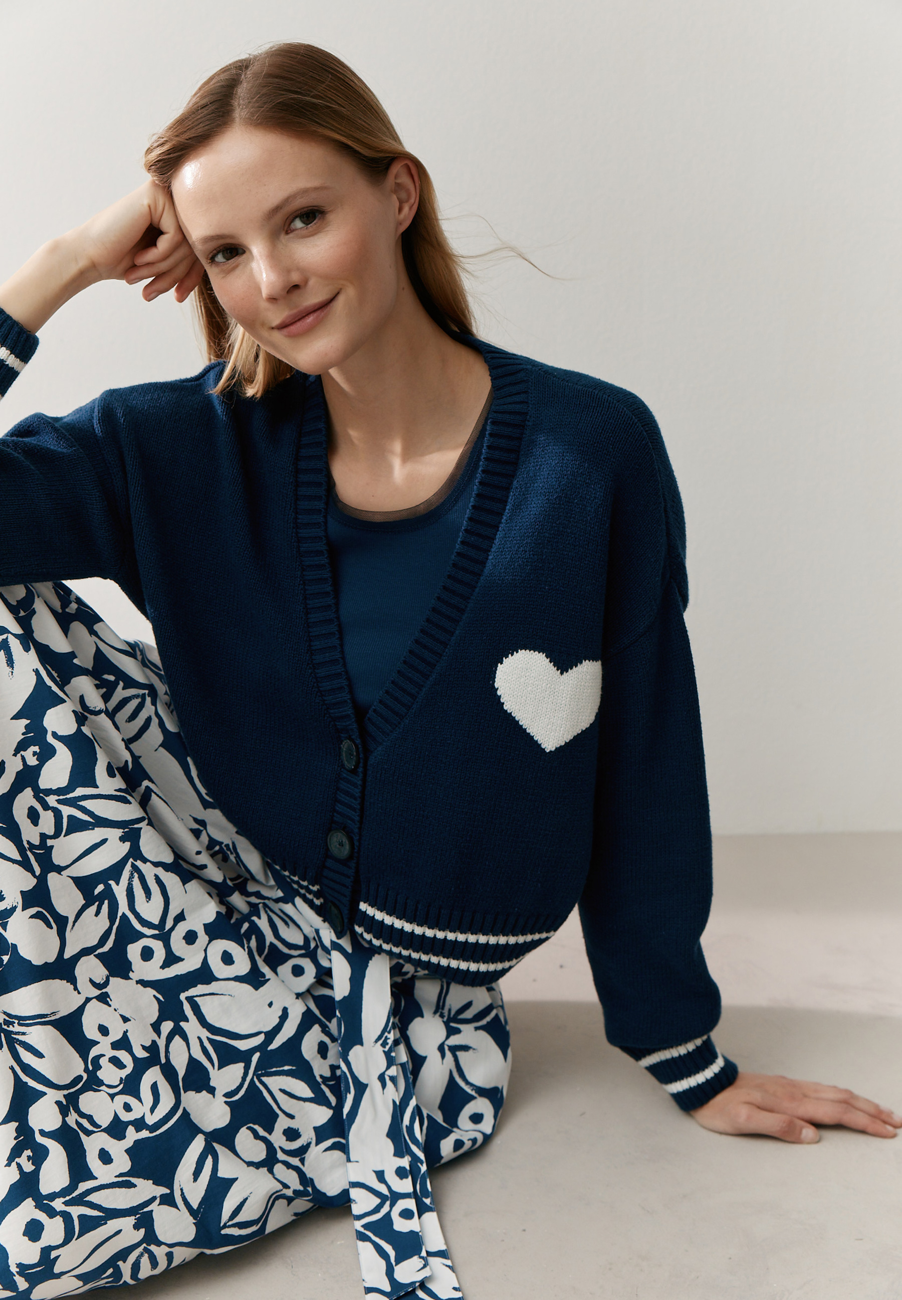 Eine Frau mit MORE & MORE Strickjacke mit Herz sitzt lächelnd am Boden.