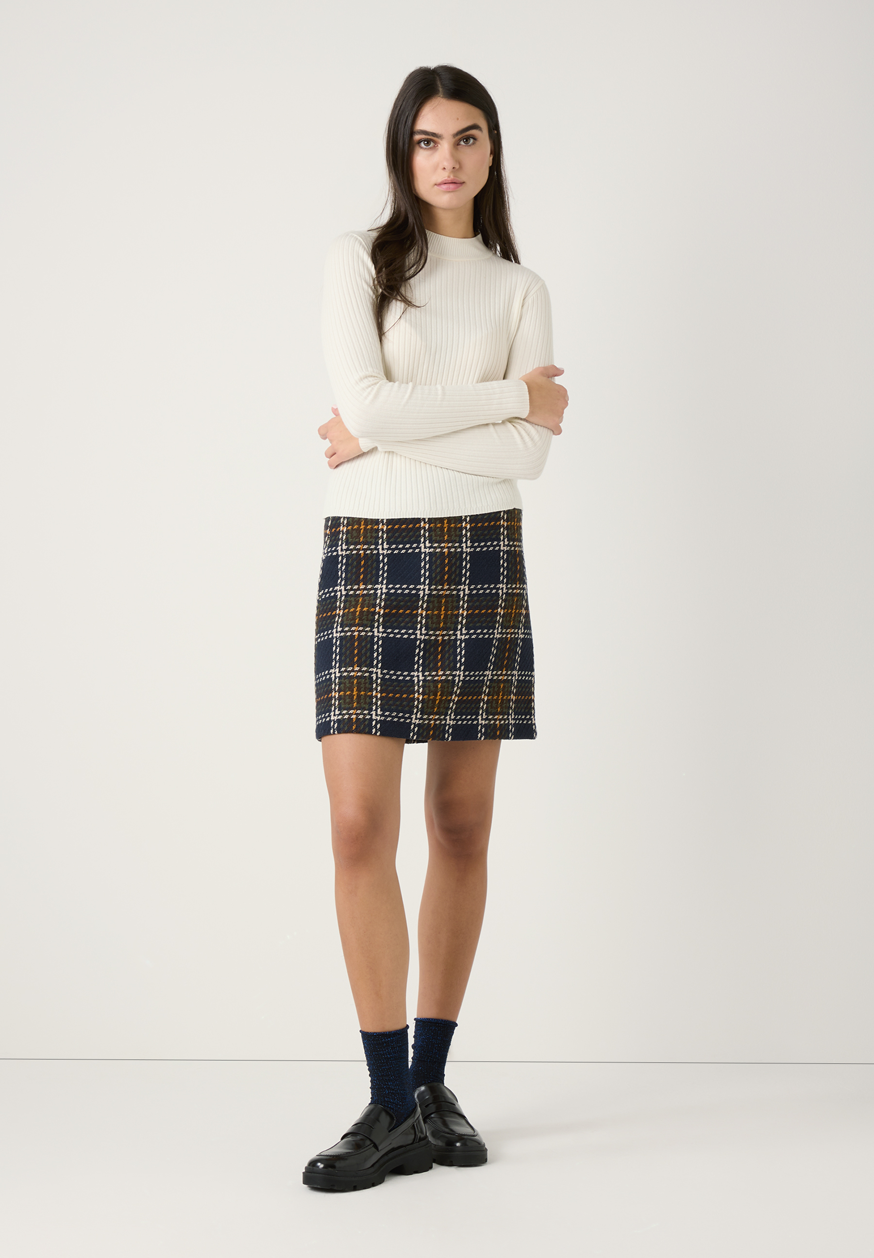 Eine Frau steht vor einem weißen Hintergrund und trägt einen MORE & MORE Strickpullover, einen karierten Minirock, Socken und Schuhe. Eine Frau steht vor einem weißen Hintergrund und trägt einen MORE & MORE Strickpullover, einen karierten Minirock, Socken und Schuhe.