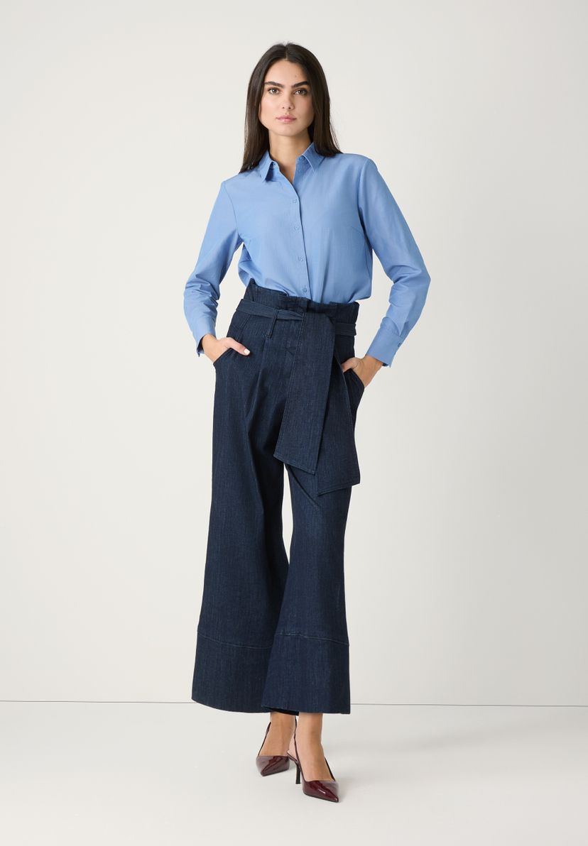 Eine Frau in einer blauen MORE & MORE Bluse und dunklen, weiten Hosen posiert vor einem weißen Hintergrund. Eine Frau in einer blauen MORE & MORE Bluse und dunklen, weiten Hosen posiert vor einem weißen Hintergrund.