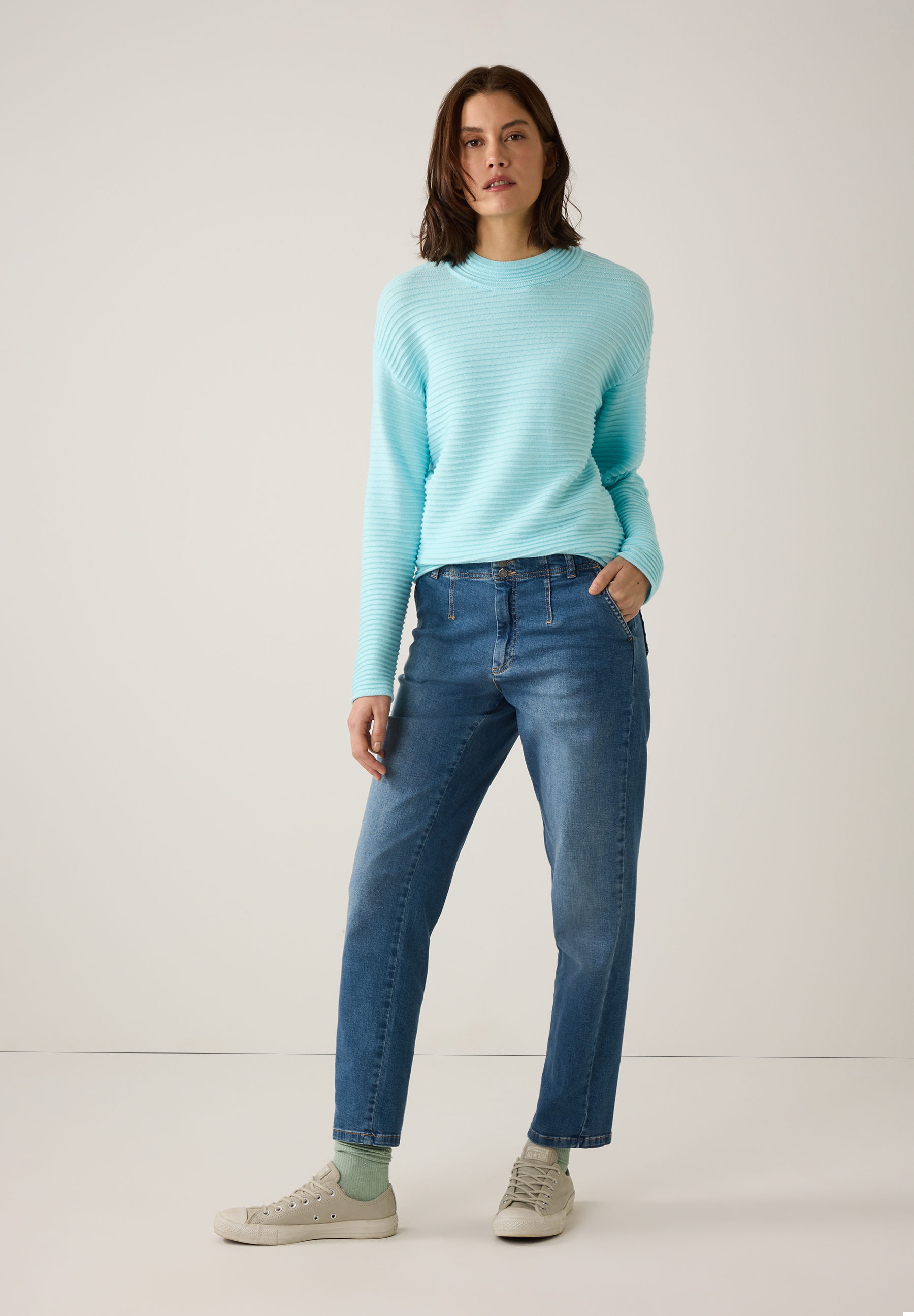 Eine Frau mit MORE & MORE Strickpullover, Jeans und Turnschuhen steht vor einem weißen Hintergrund. Eine Frau mit MORE & MORE Strickpullover, Jeans und Turnschuhen steht vor einem weißen Hintergrund.