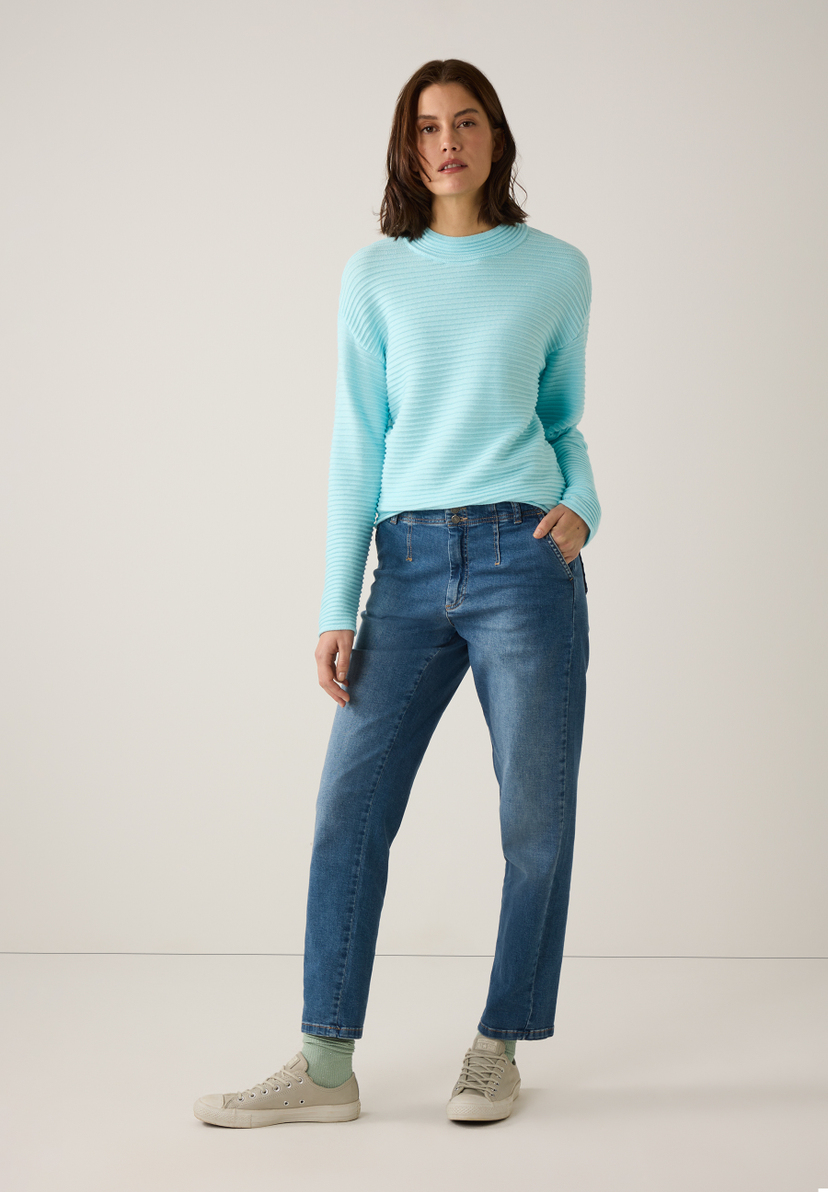 Eine Frau mit MORE & MORE Strickpullover, Jeans und Turnschuhen steht vor einem weißen Hintergrund. Eine Frau mit MORE & MORE Strickpullover, Jeans und Turnschuhen steht vor einem weißen Hintergrund.