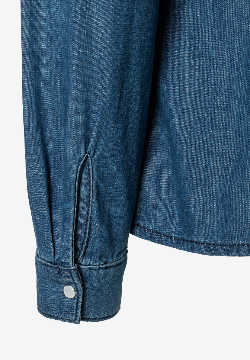 Nahaufnahme vom Ärmel einer MORE & MORE Bluse aus blauem Denim-Stoff mit Manschette und silbernem Knopf. Nahaufnahme vom Ärmel einer MORE & MORE Bluse aus blauem Denim-Stoff mit Manschette und silbernem Knopf.