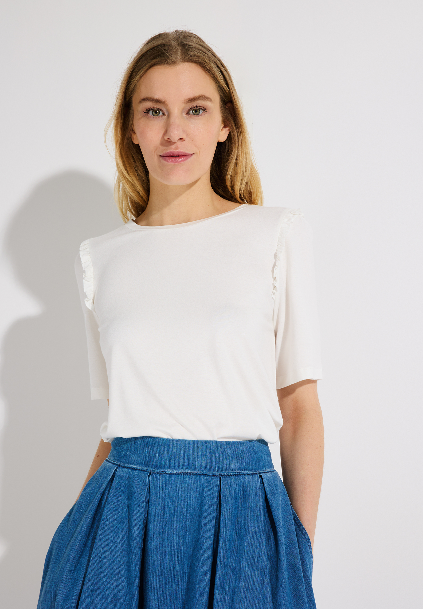 Eine Frau mit blonden Haaren trägt ein weißes MORE & MORE Oberteil und einen blauen Rock vor einem weißen Hintergrund. Eine Frau mit blonden Haaren trägt ein weißes MORE & MORE Oberteil und einen blauen Rock vor einem weißen Hintergrund.