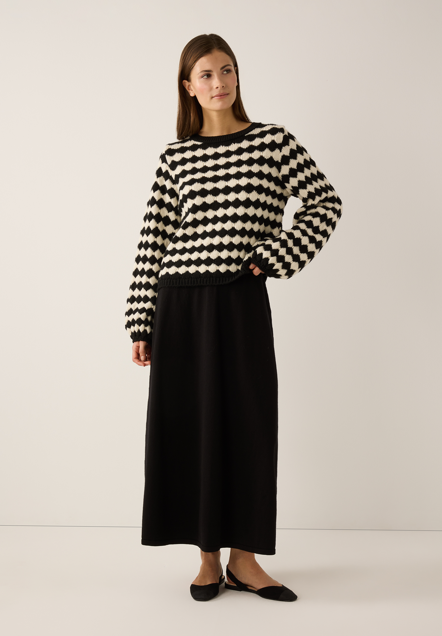 Eine Frau trägt einen MORE & MORE Strickpullover mit schwarz-weißem Muster, einen schwarzen Rock und passende Schuhe. Eine Frau trägt einen MORE & MORE Strickpullover mit schwarz-weißem Muster, einen schwarzen Rock und passende Schuhe.