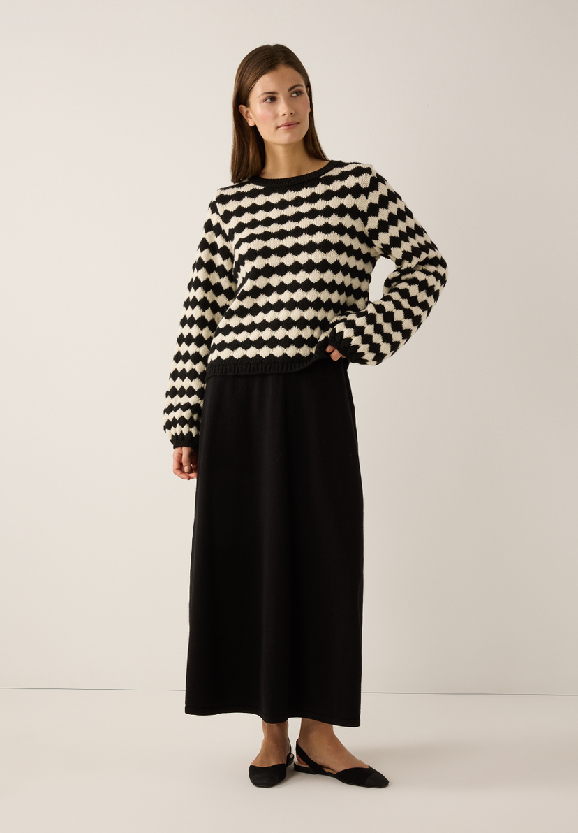 Eine Frau trägt einen MORE & MORE Strickpullover mit schwarz-weißem Muster, einen schwarzen Rock und passende Schuhe. Eine Frau trägt einen MORE & MORE Strickpullover mit schwarz-weißem Muster, einen schwarzen Rock und passende Schuhe.
