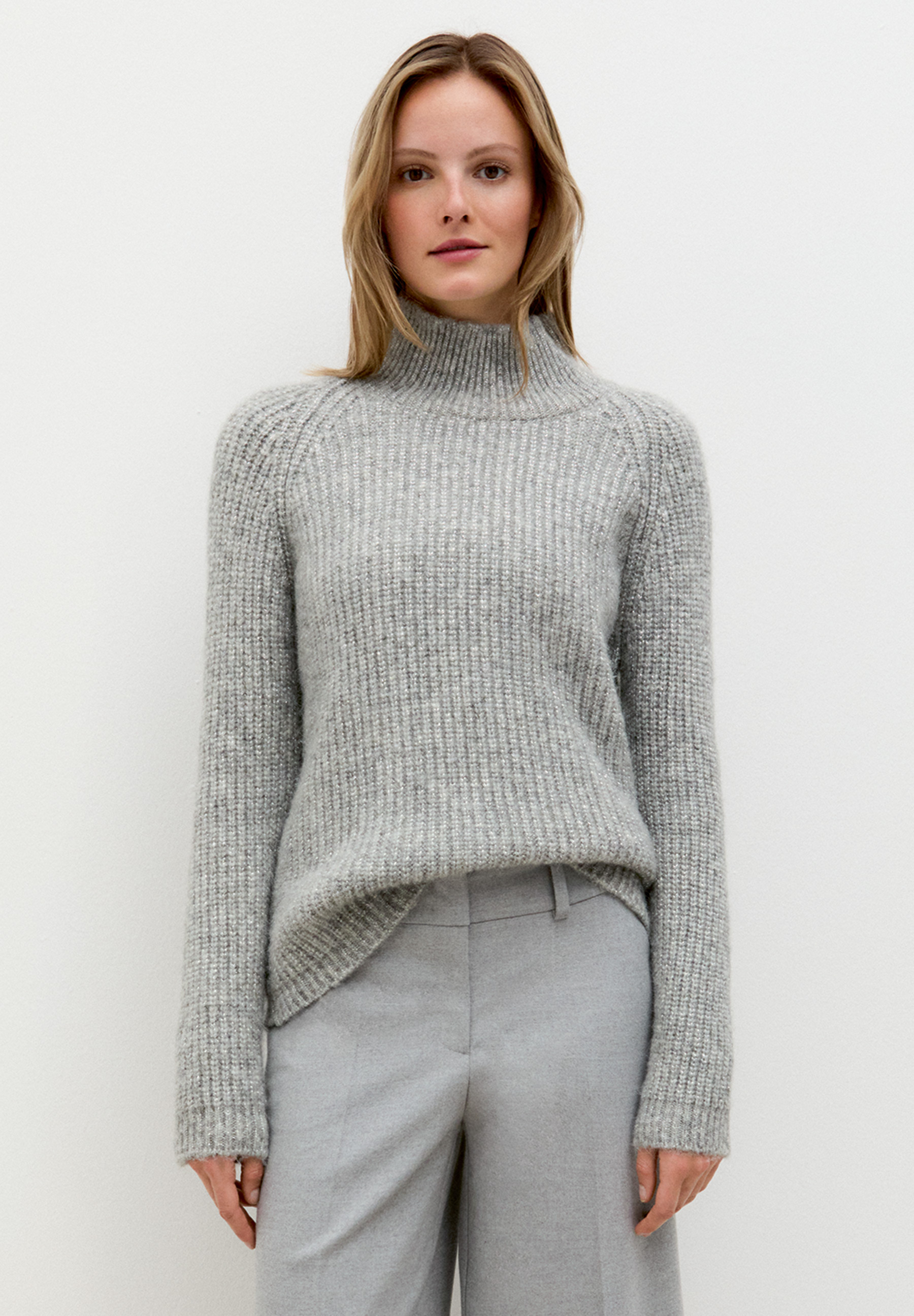 Eine Frau trägt einen grauen MORE & MORE Strickpullover, kombiniert mit einer grauen Hose vor einem weißen Hintergrund.