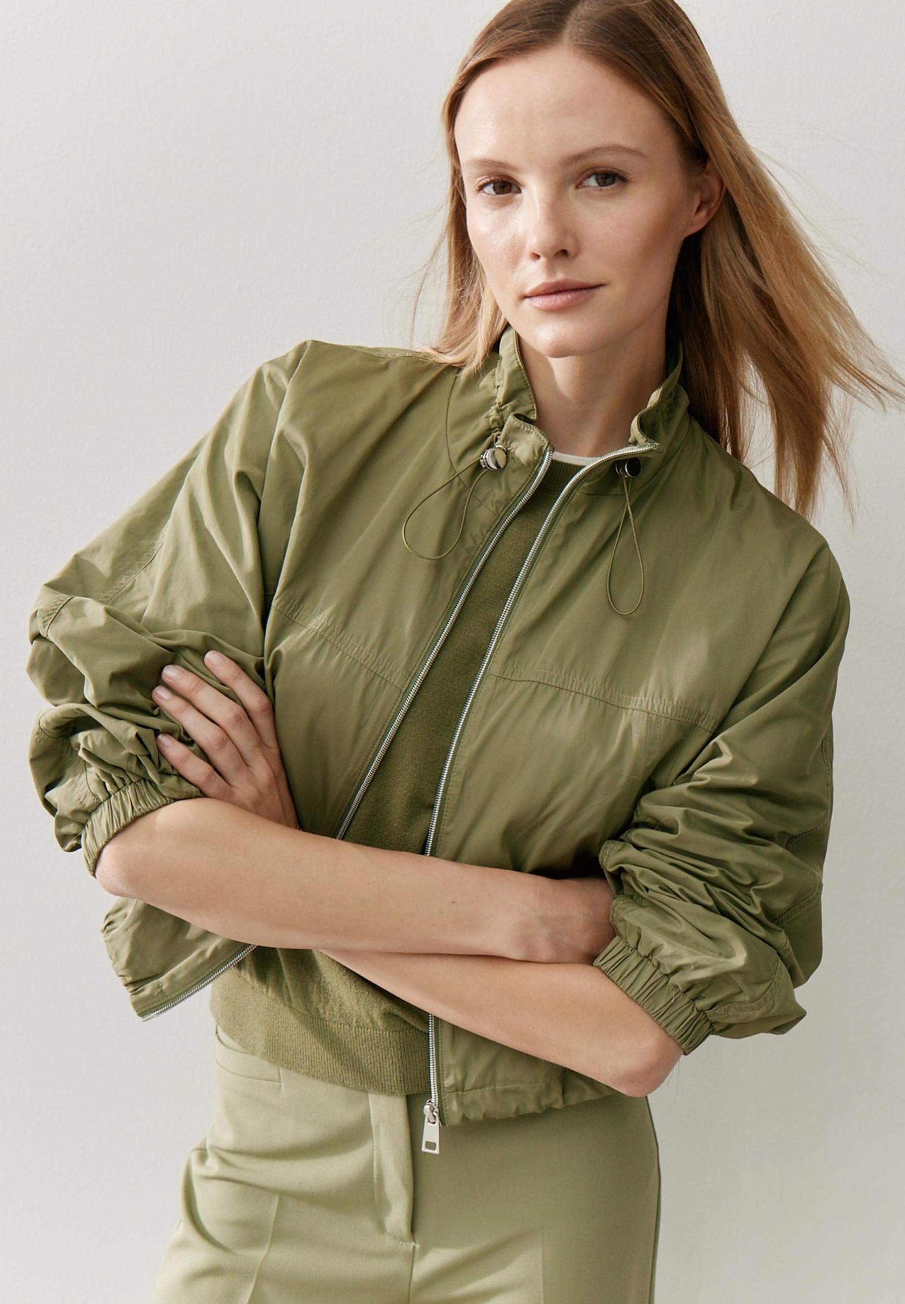 Eine Frau mit verschränkten Armen trägt einen olivgrünen MORE & MORE Blazer mit glänzender Oberfläche. Eine Frau mit verschränkten Armen trägt einen olivgrünen MORE & MORE Blazer mit glänzender Oberfläche.