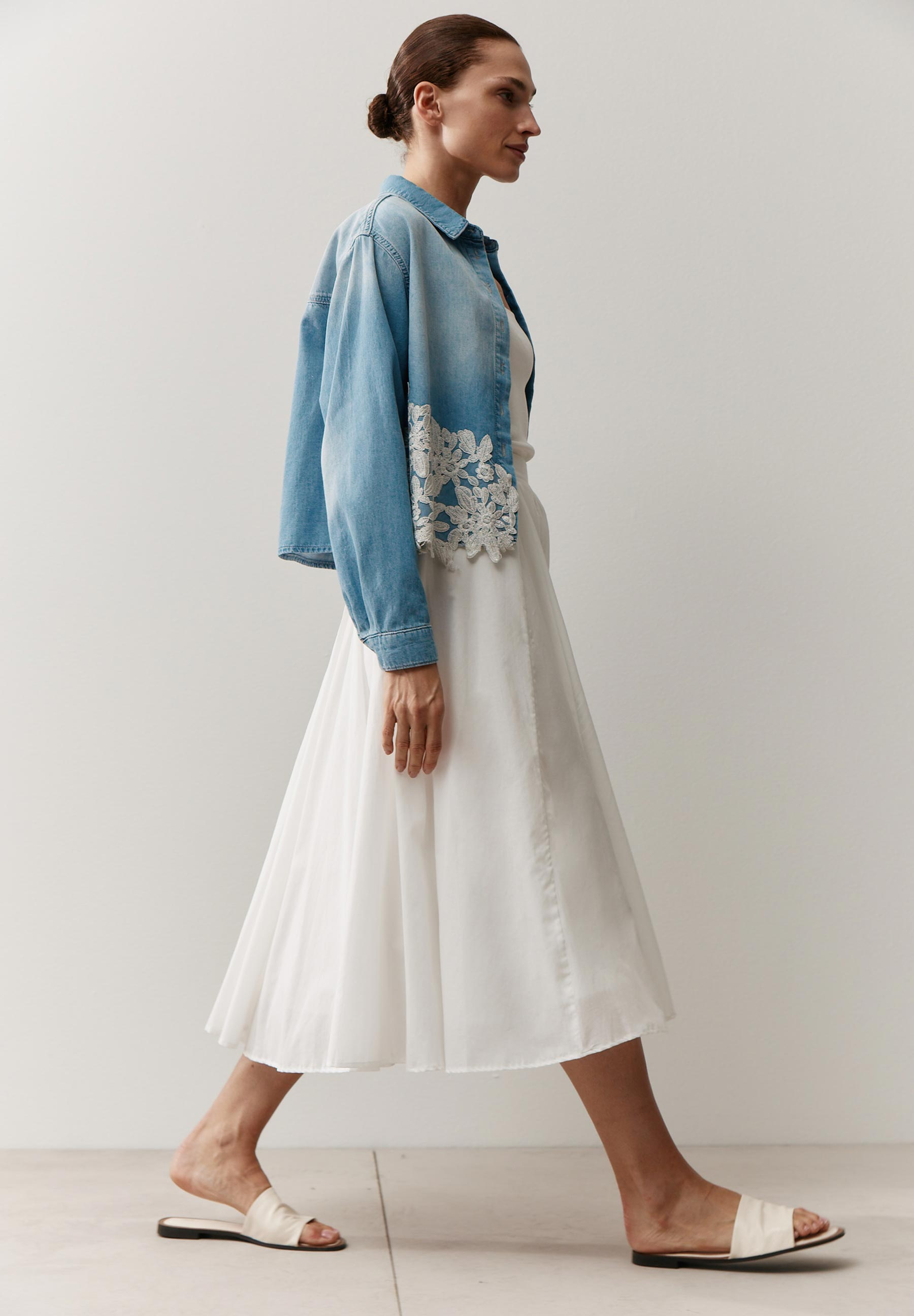 Eine Frau in einem MORE & MORE Oberteil aus Denim mit weißem Kleid und Sandalen.