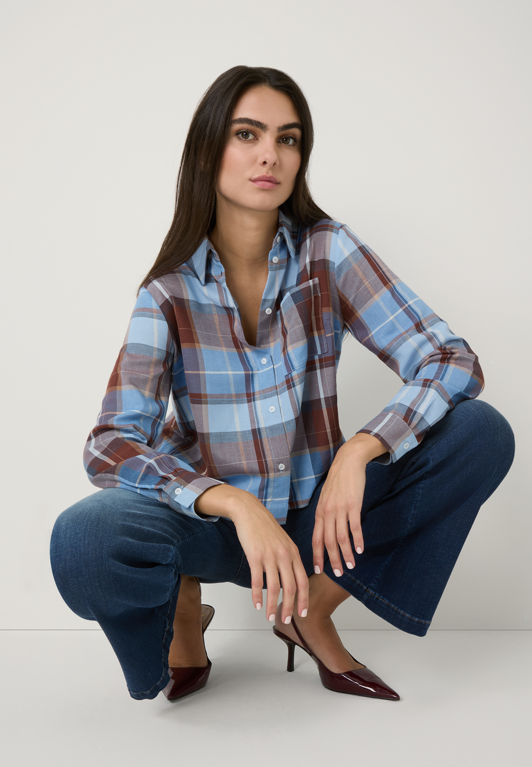 Eine Frau in MORE & MORE Jeans hockt, mit kariertem Hemd und roten Schuhen vor neutralem Hintergrund. Eine Frau in MORE & MORE Jeans hockt, mit kariertem Hemd und roten Schuhen vor neutralem Hintergrund.