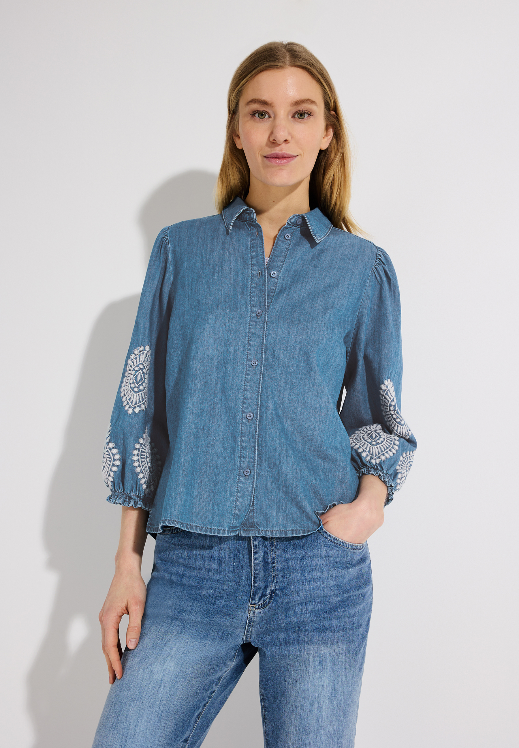 Eine blonde Frau, die ein Jeanshemd von MORE & MORE mit Stickerei auf den Ärmeln und Jeans trägt.