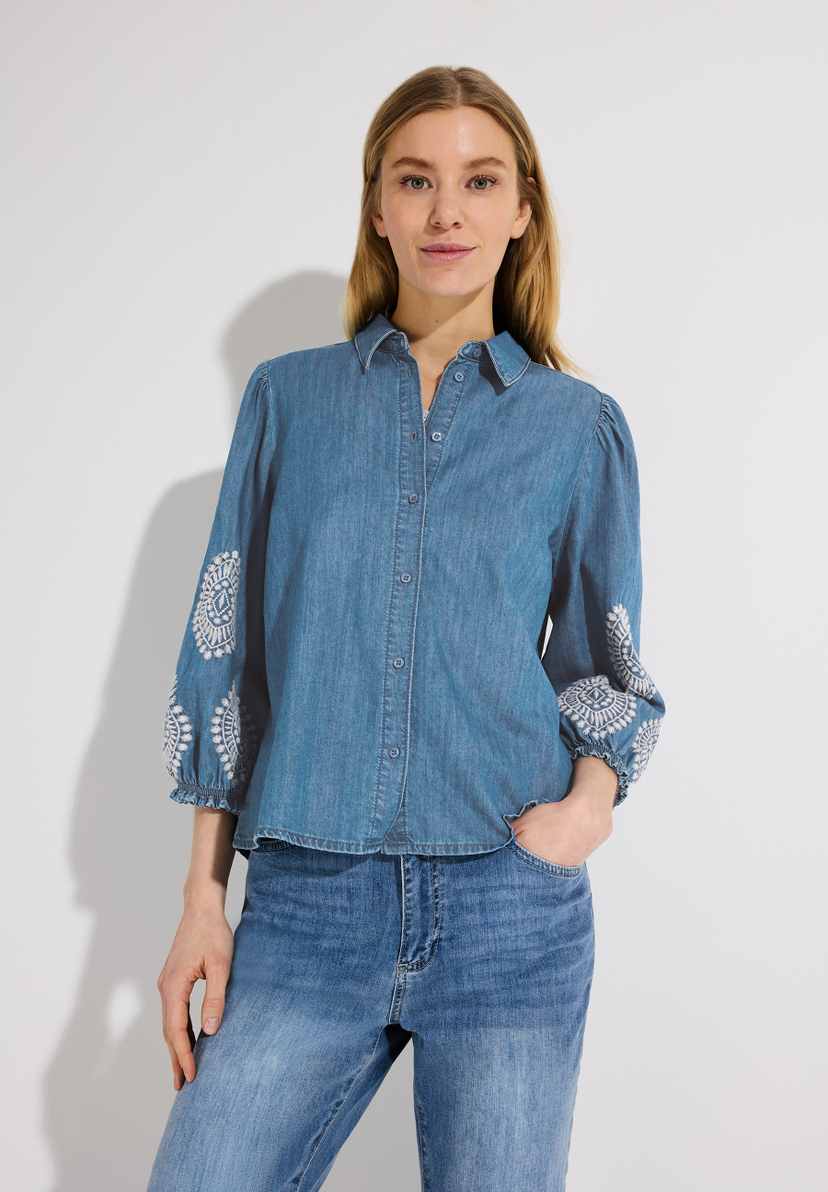 Eine blonde Frau, die ein Jeanshemd von MORE & MORE mit Stickerei auf den Ärmeln und Jeans trägt. Eine blonde Frau, die ein Jeanshemd von MORE & MORE mit Stickerei auf den Ärmeln und Jeans trägt.