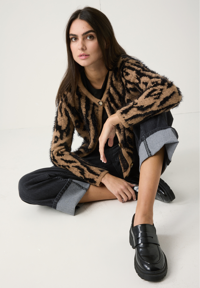 Eine Frau sitzt im MORE & MORE Strick-Cardigan mit auffälligem Muster und Jeans auf dem Boden. Eine Frau sitzt im MORE & MORE Strick-Cardigan mit auffälligem Muster und Jeans auf dem Boden.