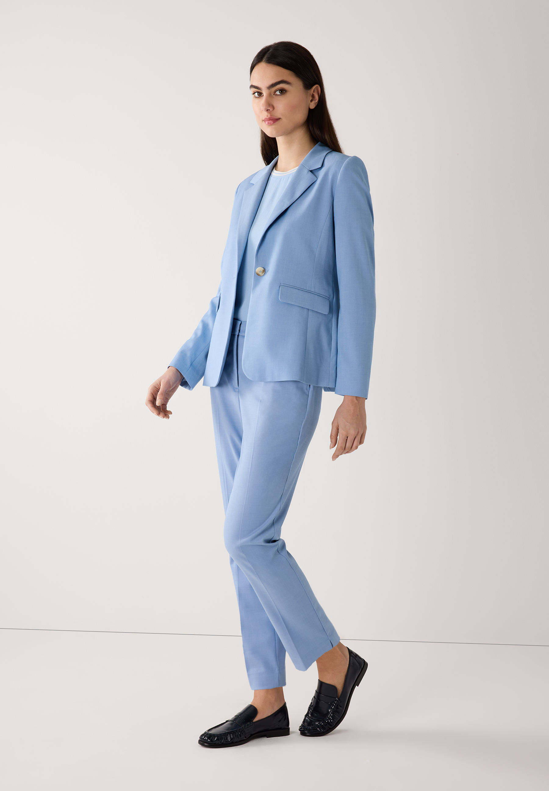 Eine Frau in einem hellblauen MORE & MORE Blazer mit passender Hose und schwarzen Loafers vor einem weißen Hintergrund. Eine Frau in einem hellblauen MORE & MORE Blazer mit passender Hose und schwarzen Loafers vor einem weißen Hintergrund.