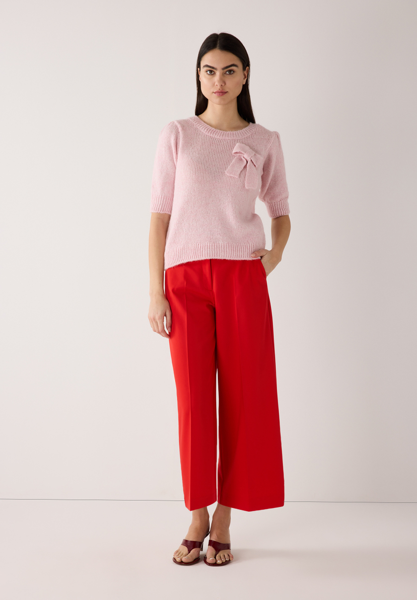 Eine Frau trägt einen MORE & MORE Strickpullover in Rosa mit Schleife und eine rote Hose. Eine Frau trägt einen MORE & MORE Strickpullover in Rosa mit Schleife und eine rote Hose.