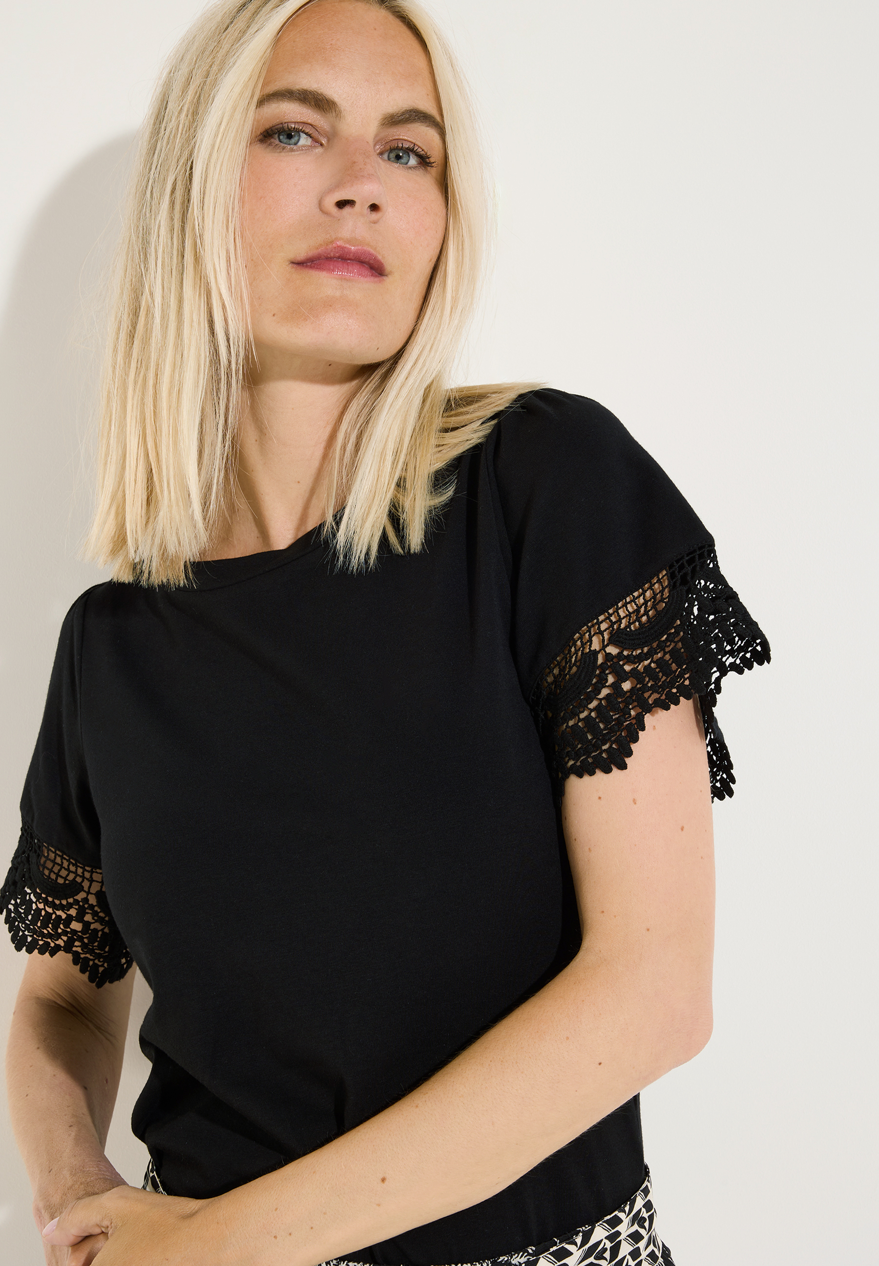 MORE & MORE Damenshirt in Schwarz mit kurzen Ärmeln und Häkelspitze. Halbporträt einer blonden Frau mit blauen Augen.
