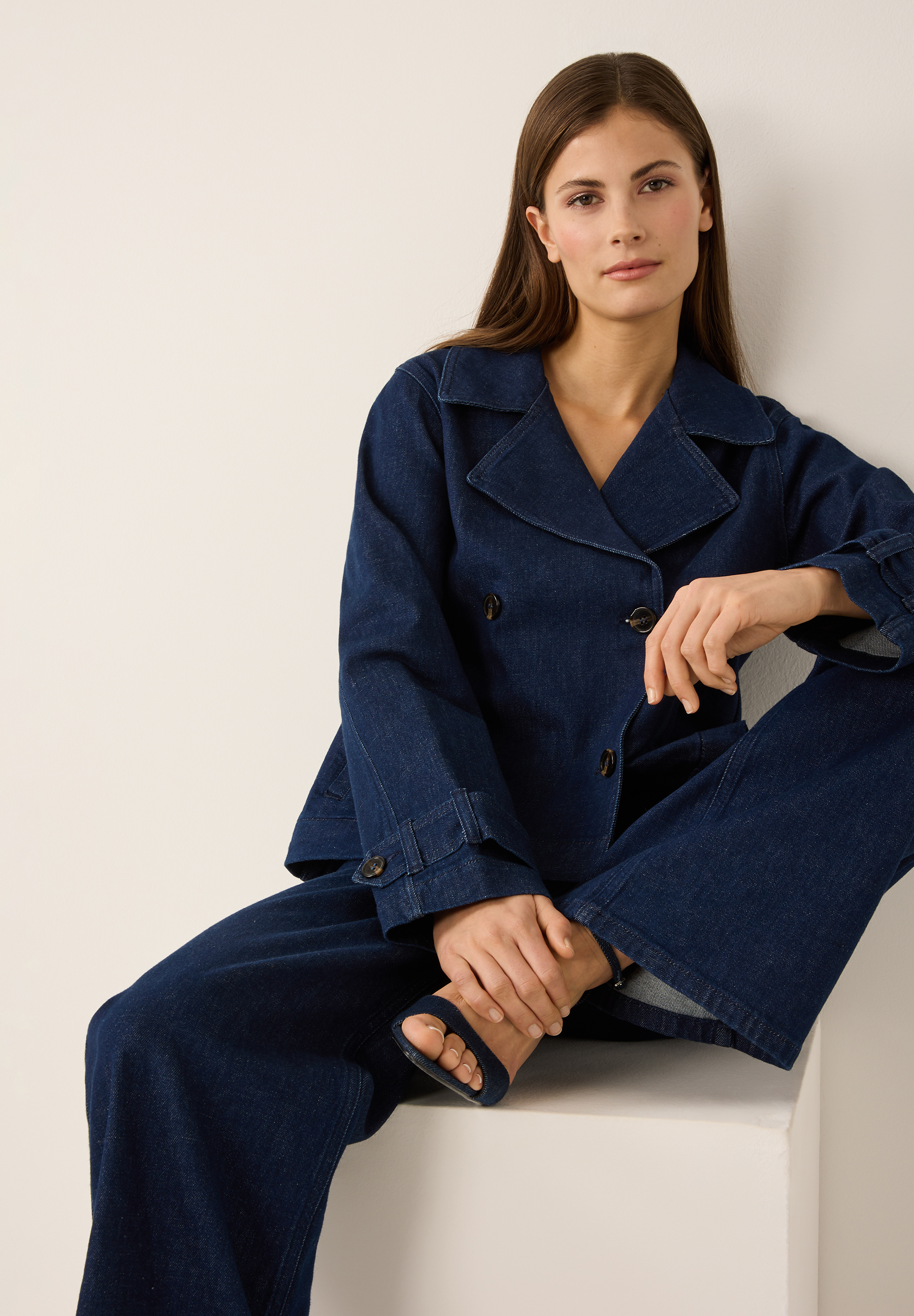 Eine Frau in einem MORE & MORE Denim-Outfit sitzt lässig vor einer hellen Wand. Eine Frau in einem MORE & MORE Denim-Outfit sitzt lässig vor einer hellen Wand.