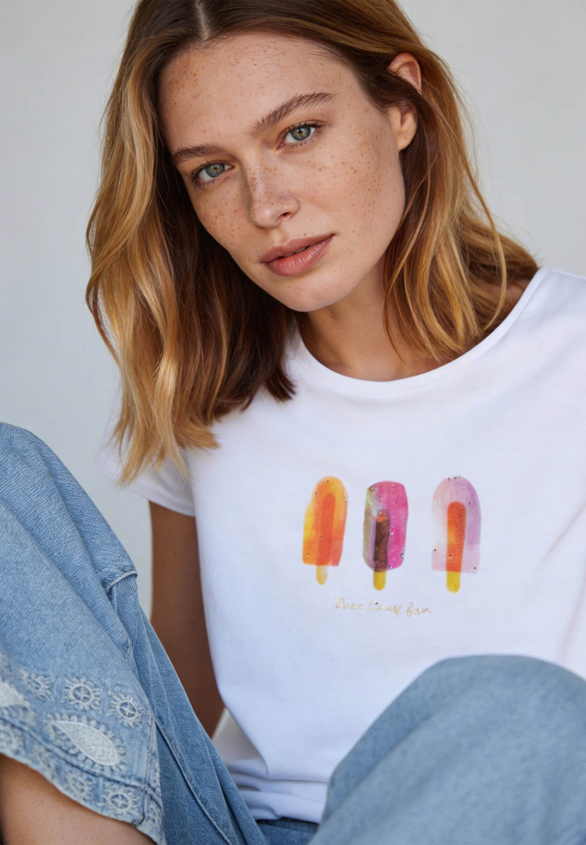 Eine Frau in einem weißen MORE & MORE T-Shirt mit Eis-Motiven und Jeans. Eine Frau in einem weißen MORE & MORE T-Shirt mit Eis-Motiven und Jeans.