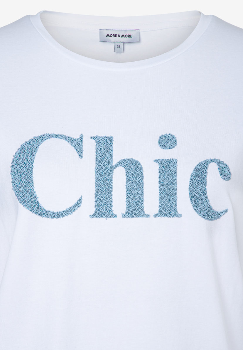 Ein weißes MORE & MORE T-Shirt mit blauem "Chic"-Schriftzug aus kleinen Kugeln. Ein weißes MORE & MORE T-Shirt mit blauem "Chic"-Schriftzug aus kleinen Kugeln.