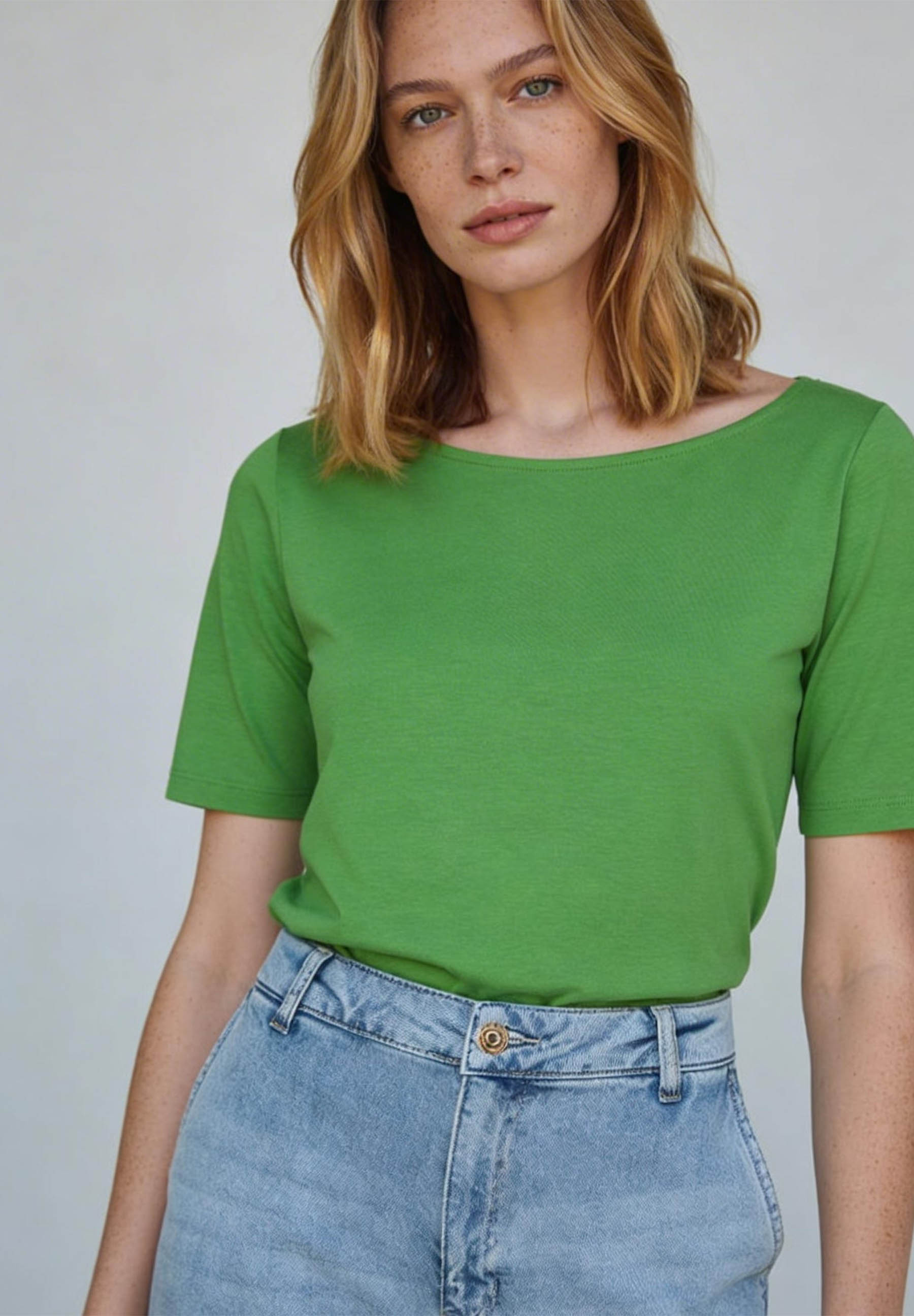 Eine Frau trägt ein grünes Jersey-Top von MORE & MORE und Jeans. Das Shirt hat einen U-Boot-Ausschnitt und kurze Ärmel.
