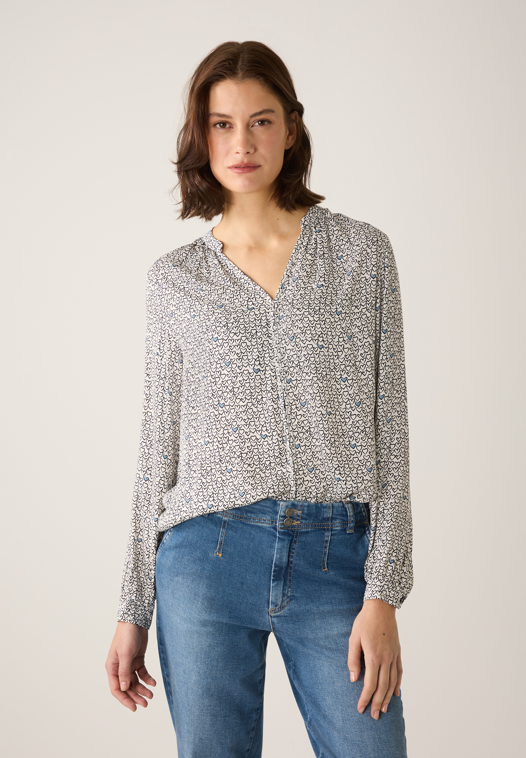 Eine Frau trägt eine MORE & MORE Bluse mit Herzmuster und blaue Jeans.